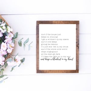 May include: A framed wooden sign with the text "And if the house just keeps on winning...and keep a bluebird in my heart" in black font. The sign has a white background and a dark brown frame. Floral arrangement with pink flowers and green leaves.