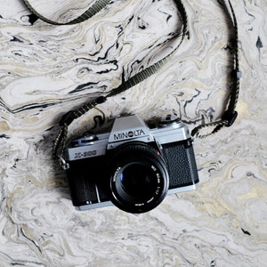 May include: A vintage Minolta X-300 camera with a black lens and a green and black strap rests on a marbled surface. The camera body is silver and black, with the Minolta logo visible. The strap is woven.