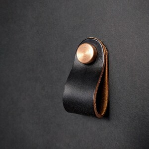 May include: A black leather pull with a copper-colored rivet. The pull is looped and attached to a dark surface. The leather has a textured appearance, and the rivet is circular.