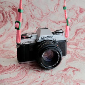 Könnte beinhalten: Eine silber-schwarze Minolta X-300 35mm Filmkamera mit einem Minolta MD 50mm 1:1.7 Objektiv. Die Kamera verfügt über einen leuchtend pinken, gewebten Trageriemen mit grünen Kunststoff-Verstellschnallen und Schiebern an den seitlichen Ösen.