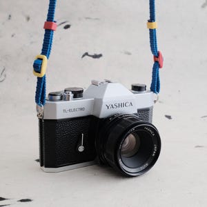 May include: A silver and black Yashica TL-Electro 35mm film camera with a black lens. The camera features a blue fabric neck strap with adjustable yellow and red plastic sliders. The lens is marked with Yashica Japan Auto text.
