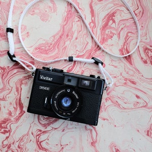 May include: A black Vivitar 35EE automatic camera with a white camera strap on a pink and white marbled background.
