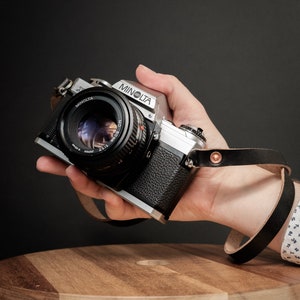 May include: A silver Minolta camera with a black lens is held in a hand. The camera is attached to a black leather wrist strap. The camera is sitting on a wooden stool.