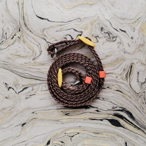 May include: A coiled brown and tan rope leash with yellow and orange accents. The leash has a braided texture and includes metal rings. The background features a marbled pattern with gold and black veining.