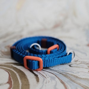 May include: A coiled blue nylon strap with orange and silver hardware. The strap features an orange buckle and two orange loops. The background is a marbled pattern in shades of white, brown, and gray.