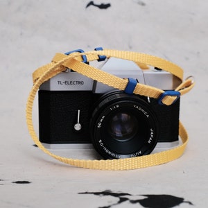 Può includere: Una macchina fotografica a pellicola da 35 mm nera e argento con un cinturino per fotocamera giallo e blu. L'obiettivo della fotocamera dice "Yashica Auto 50mm 1:1.9 Japan" e "Ashinon-DS". Il corpo della fotocamera dice "TL-Electro".