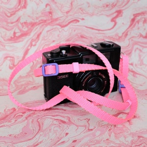May include: A black Vivitar 35EE film camera with a bright pink strap and purple accents. The camera is set against a pink and white marbled background. The strap is looped around the camera.