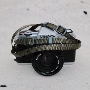 Könnte beinhalten: Eine silberne Minolta Kamera mit einem schwarzen Objektiv und einem grünen Gurt. Das Objektiv trägt die Aufschrift "Minolta MD 50mm 1:1.7 Japan".