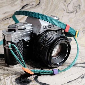 May include: A silver Minolta X-300 camera with a black lens and a teal camera strap with orange and purple accents.