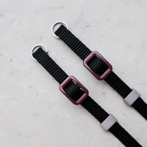 Peut inclure: Deux sangles en nylon noir avec des anneaux en métal argenté et des boucles en plastique bordeaux. Les sangles ont des curseurs en plastique gris. Image en gros plan sur fond blanc texturé.
