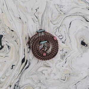 May include: A coiled brown rope with light blue and pink accents. The rope is adorned with two light blue oval-shaped beads and two pink square beads. The background features a marbled pattern in shades of white, black, and gold.