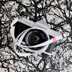 May include: A silver and black Yashica TL-Electro 35mm film camera with a 50mm 1:1.9 lens. The camera features a white woven neck strap with small red and brown plastic accents wrapped around the lens barrel, displaying the brand name Yashica.