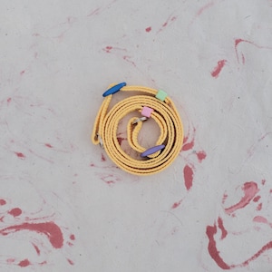 Pode incluir: Um cinto amarelo enrolado com detalhes coloridos. O cinto apresenta elementos decorativos azuis, verdes, rosa e roxos. O fundo é uma superfície texturizada esbranquiçada com marmoreio rosa e vermelho.