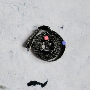 May include: A coiled lanyard with a black and white patterned cord. The lanyard features a black clasp, a key ring, and two small, square beads in pink and purple. The background is a textured, off-white surface.
