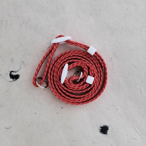 May include: A red braided camera strap with white plastic adjusters. The strap is coiled up on a white surface.