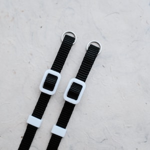 May include: Two black camera straps with white plastic adjustable buckles and silver metal rings.