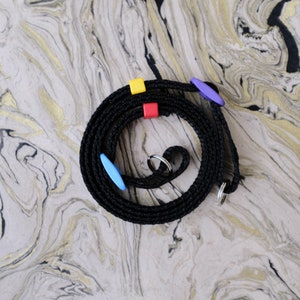 May include: Black camera strap with colorful plastic accents. The strap is coiled and has two metal rings.