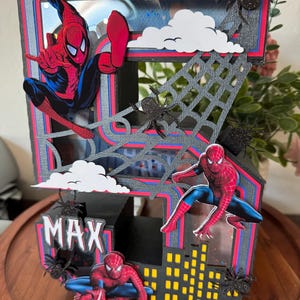 Op de afbeelding: Een handgemaakt meerlagig papieren kunstwerk met Spider-Man. Het ontwerp omvat een stadsgezicht, spinnenwebben en de naam "MAX". Het kleurenschema is rood, blauw, zwart en geel, met witte wolkenaccenten en zwarte spinnendecoraties.