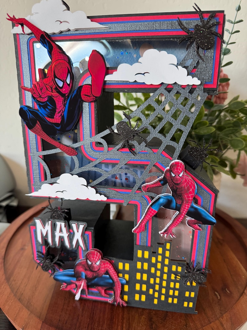 Op de afbeelding: Een handgemaakt meerlagig papieren kunstwerk met Spider-Man. Het ontwerp omvat een stadsgezicht, spinnenwebben en de naam "MAX". Het kleurenschema is rood, blauw, zwart en geel, met witte wolkenaccenten en zwarte spinnendecoraties.