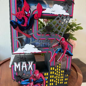 Op de afbeelding: Handgemaakte verjaardagskaart met Spider-Man. De kaart is zwart, rood en blauw, met de naam "MAX" erop. Het bevat Spider-Man-figuren, een skyline, spinnenwebben en wolkuitsparingen.