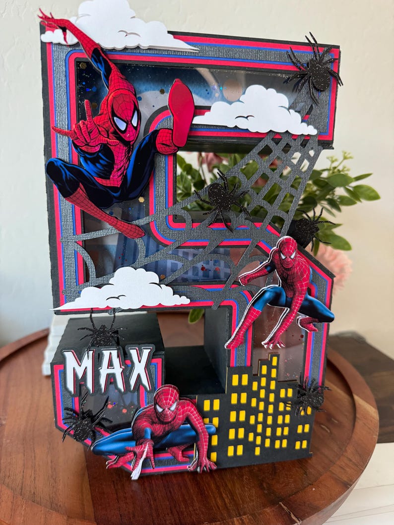 Op de afbeelding: Handgemaakte verjaardagskaart met Spider-Man. De kaart is zwart, rood en blauw, met de naam "MAX" erop. Het bevat Spider-Man-figuren, een skyline, spinnenwebben en wolkuitsparingen.