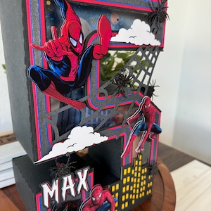 Op de afbeelding: Een meerlagige, zwart-rode decoratieve doos met Spider-Man. De doos heeft een stadsgezichtontwerp met de naam "MAX" en spinnenwebaccenten. Spider-Man wordt afgebeeld in verschillende actieposes, met witte wolken en zwarte spinnen.