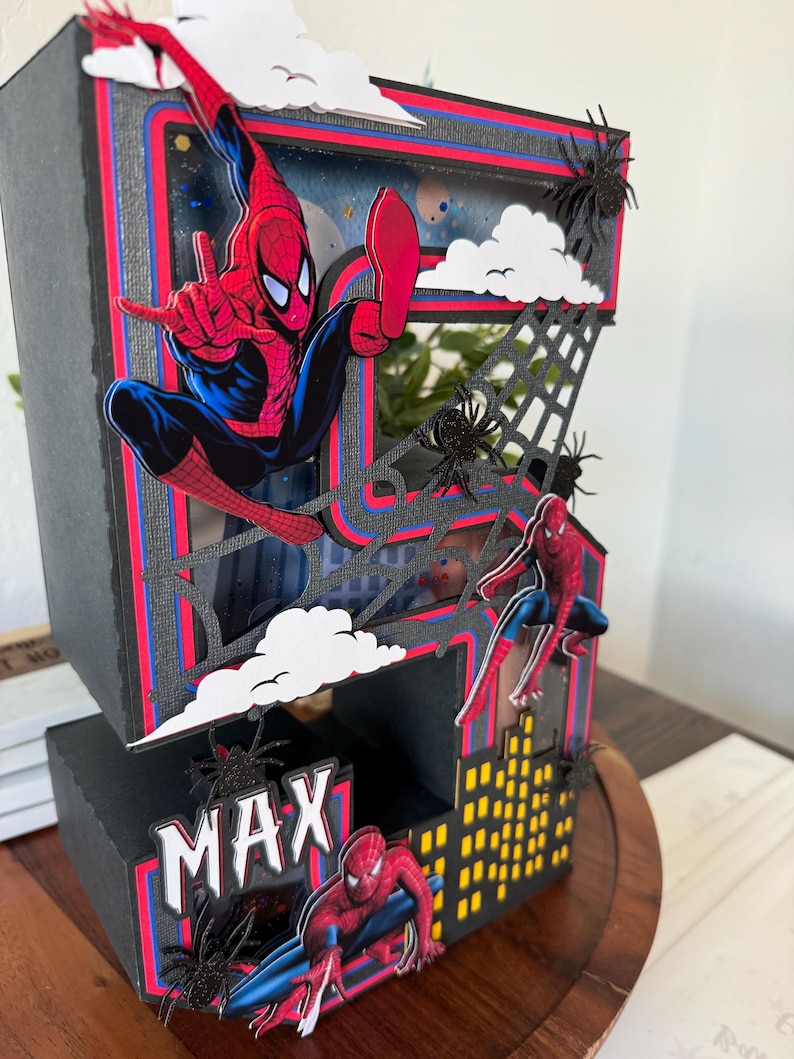 Op de afbeelding: Een meerlagige, zwart-rode decoratieve doos met Spider-Man. De doos heeft een stadsgezichtontwerp met de naam "MAX" en spinnenwebaccenten. Spider-Man wordt afgebeeld in verschillende actieposes, met witte wolken en zwarte spinnen.
