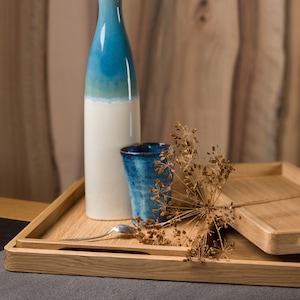 Peut inclure: Nature morte avec une bouteille en céramique bleue et blanche, une petite tasse bleue et un plateau en bois. Une cuillère en argent et des brins de plantes séchées ajoutent des détails. Sur fond gris et bois.