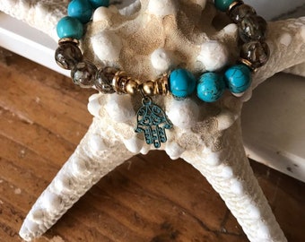 May include: A turquoise and brown beaded bracelet with a gold charm in the shape of a hamsa hand. The bracelet is on a white starfish.
