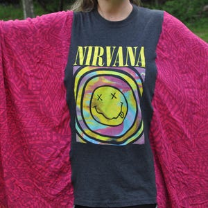 Könnte beinhalten: Ein dunkelgraues T-Shirt mit dem Wort "NIRVANA" in Gelb, über einem bunten Smiley-Design. Das Shirt wird mit einem hellrosa gemusterten Schal kombiniert.