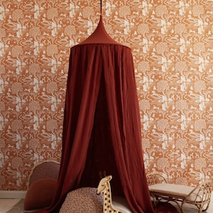 Può includere: Una tenda a baldacchino in tessuto rosso pende dal soffitto in una stanza con carta da parati a motivi. All'interno della tenda, c'è una luce notturna a forma di giraffa in legno, un cuscino e un tappeto intrecciato.