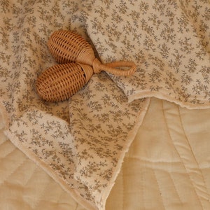 May include: A woven rattan baby rattle lies on a cream-coloured blanket with a floral pattern. The blanket features a delicate design of small grey flowers. The blanket is placed on a quilted cream-coloured surface.