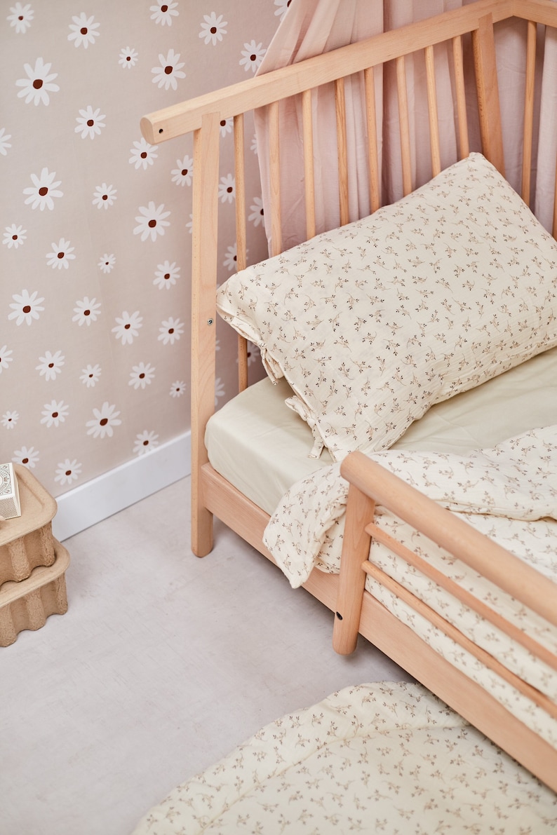 K&ouml;nnte beinhalten: Ein Holzbettchen mit einem wei&szlig;en, floral gemusterten Bettw&auml;sche-Set. Das Bettchen hat einen Baldachin und steht an einer Wand mit einem wei&szlig;en G&auml;nsebl&uuml;mchenmuster.