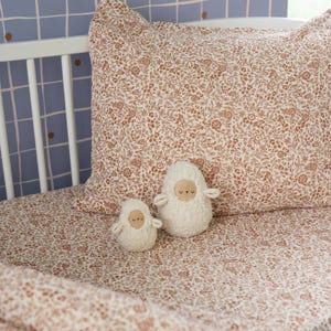 Peut inclure: Un lit bébé avec un ensemble de draps et d'oreillers à motifs floraux dans les tons crème et marron. Deux jouets moutons en peluche blancs avec des visages beiges et de petits bras sont sur le drap. L'arrière-plan présente un mur bleu avec un motif quadrillé.