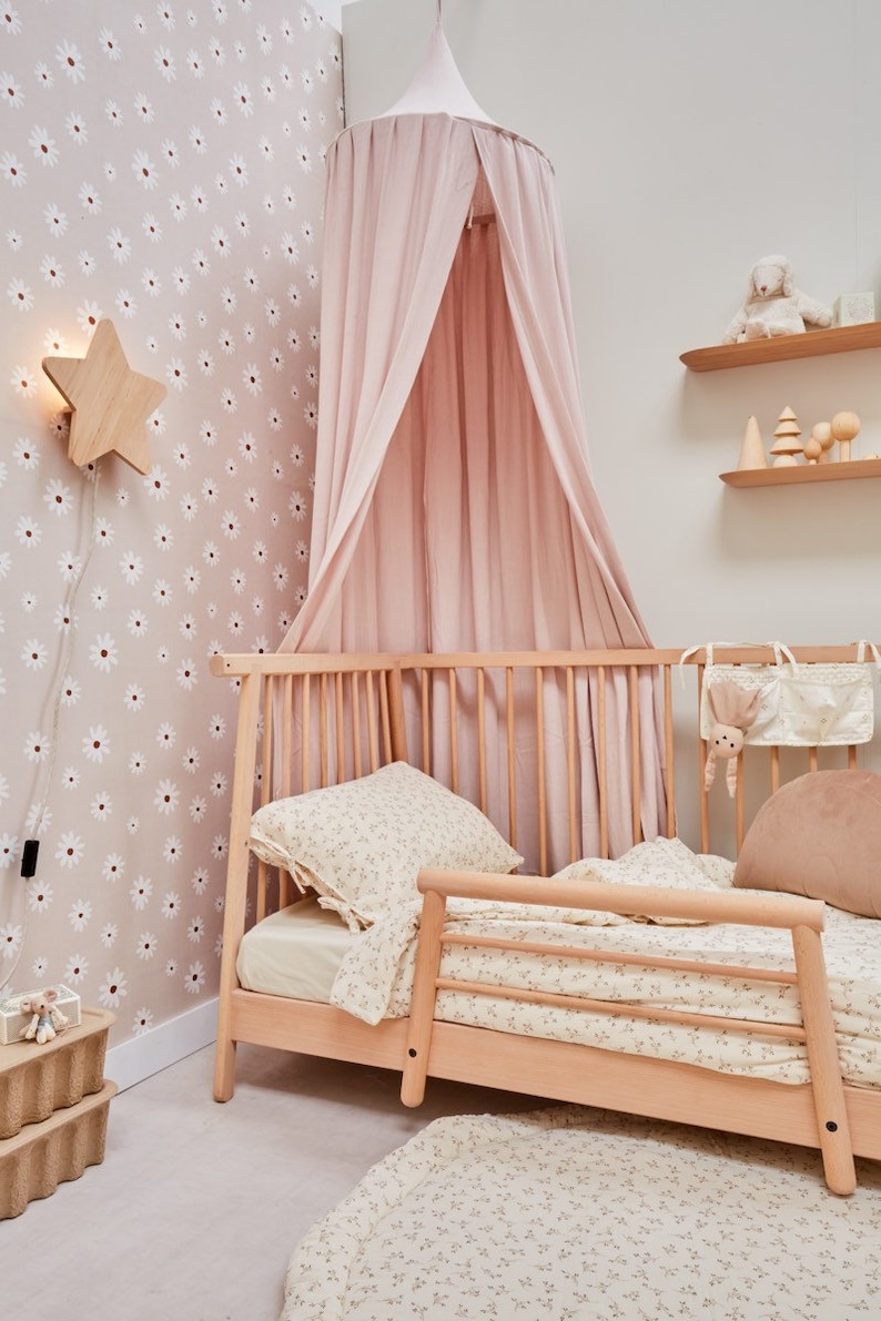 K&ouml;nnte beinhalten: Ein rosa Himmelbett mit einem Holzrahmen und Bettw&auml;sche mit Blumenmuster. Das Bett steht in einem Zimmer mit Blumenmuster-Tapete und einer sternf&ouml;rmigen Nachtischlampe aus Holz.