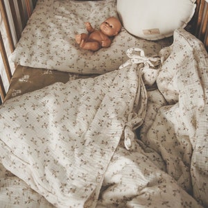 K&ouml;nnte beinhalten: Ein wei&szlig;es Babybett mit einem wei&szlig;en und braunen Bettw&auml;sche-Set mit Blumenmuster. Eine Puppe liegt auf der Bettw&auml;sche. Das Babybett hat Holzlatten und einen wei&szlig;en Baldachin.