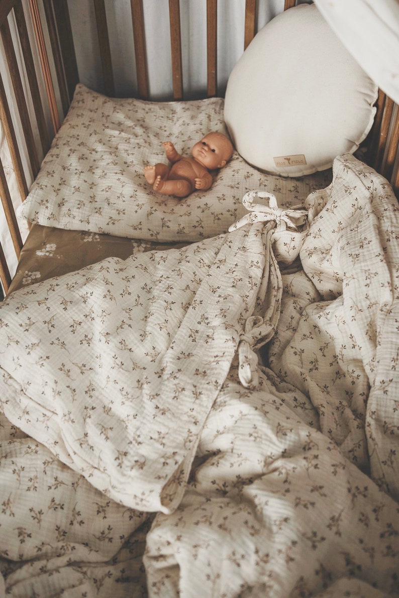 K&ouml;nnte beinhalten: Ein wei&szlig;es Babybett mit einem wei&szlig;en und braunen Bettw&auml;sche-Set mit Blumenmuster. Eine Puppe liegt auf der Bettw&auml;sche. Das Babybett hat Holzlatten und einen wei&szlig;en Baldachin.
