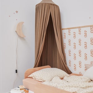 K&ouml;nnte beinhalten: Ein hellbraunes Himmelbett mit einer wei&szlig;en Bettw&auml;sche mit Blumenmuster und einem Kissen. Das Bett steht an einer Wand mit einer Blumen-Tapete und einer mondf&ouml;rmigen Nachtlampe aus Holz.