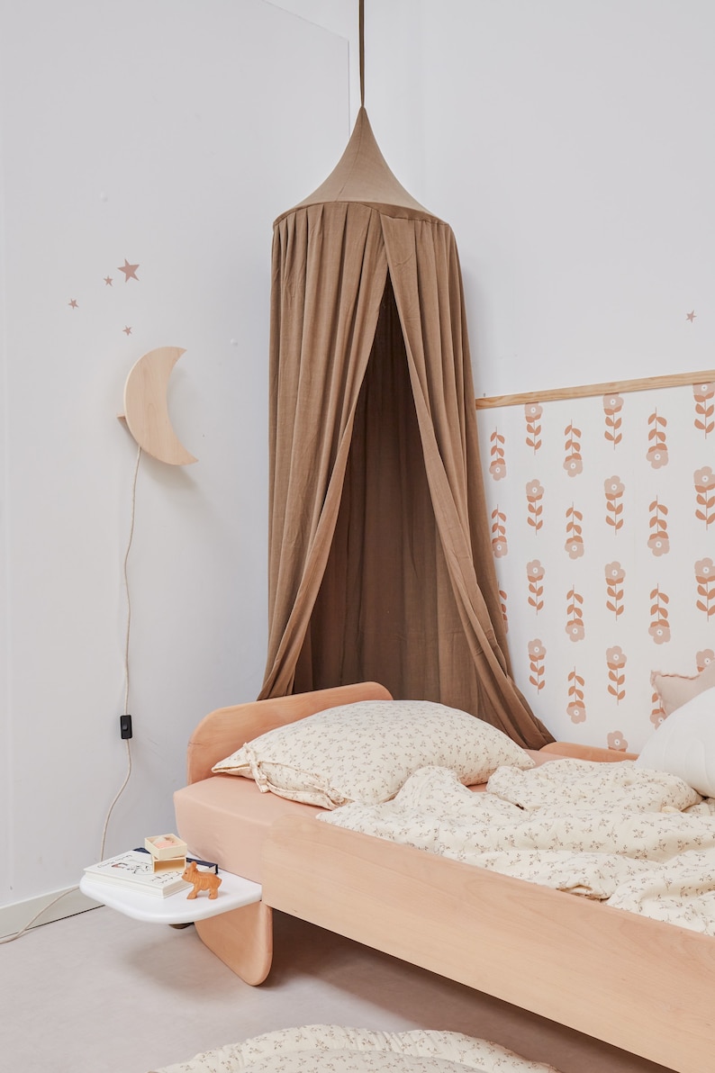 K&ouml;nnte beinhalten: Ein hellbraunes Himmelbett mit einer wei&szlig;en Bettw&auml;sche mit Blumenmuster und einem Kissen. Das Bett steht an einer Wand mit einer Blumen-Tapete und einer mondf&ouml;rmigen Nachtlampe aus Holz.
