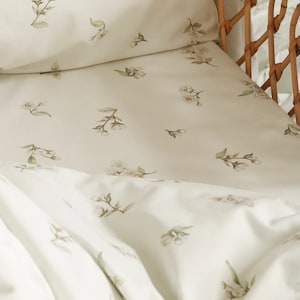 Könnte beinhalten: Weiße Bettwäsche mit einem zarten Blumenmuster aus kleinen weißen Blüten und grünen Blättern. Die Bettwäsche ist aus weichem, atmungsaktivem Stoff und perfekt für ein Kinderzimmer oder ein Gästezimmer.