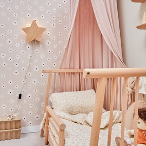 K&ouml;nnte beinhalten: Ein hellrosa Baldachinbett mit einem Holzrahmen und einer Bettw&auml;sche mit wei&szlig;em Blumenmuster. Das Bett steht in einem Zimmer mit einer wei&szlig;en Tapete mit G&auml;nsebl&uuml;mchenmuster und einer sternf&ouml;rmigen Nachtischlampe aus Holz.
