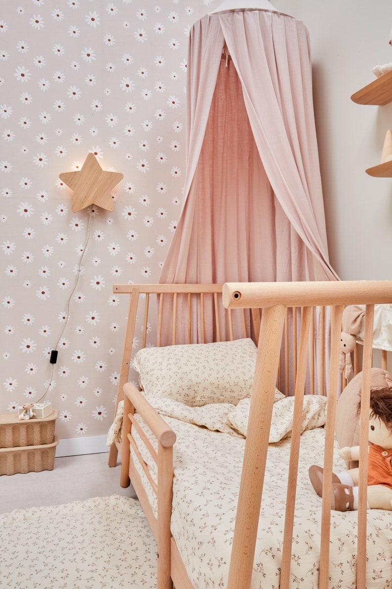 K&ouml;nnte beinhalten: Ein hellrosa Baldachinbett mit einem Holzrahmen und einer Bettw&auml;sche mit wei&szlig;em Blumenmuster. Das Bett steht in einem Zimmer mit einer wei&szlig;en Tapete mit G&auml;nsebl&uuml;mchenmuster und einer sternf&ouml;rmigen Nachtischlampe aus Holz.