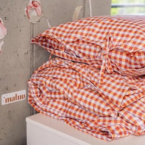 May include: A stack of orange and white gingham bedding, including a pillow and a folded blanket. The pillow has tie closures. The word "maluu" is visible on a sticker on the wall.