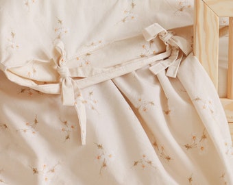 May include: Cream-colored bedding set featuring a delicate floral pattern of small white daisies with yellow centers and green stems. The pillowcase and duvet cover have matching fabric ties. The bedding is made of a soft, lightweight material.