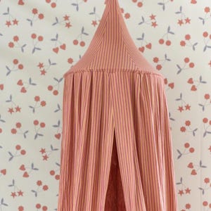 May include: A pink and yellow striped canopy with a conical top, suspended from above. The canopy features vertical stripes and a split opening. The background is a white wall with a pattern of red cherries and grey leaves.