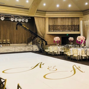 May include: A white dance floor with gold lettering that says "A & A" in a large, cursive font. The dance floor is in a large, empty ballroom with gold curtains and a white ceiling. There are tables and chairs set up around the dance floor, and there are floral centerpieces on the tables.
