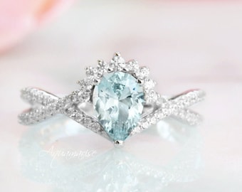 Puede incluir: Un anillo de compromiso plateado con una piedra preciosa azul claro en forma de pera en el centro. La piedra preciosa está rodeada de piedras más pequeñas y transparentes. La banda presenta un diseño entrecruzado, también adornado con pequeñas piedras transparentes. La palabra "Aquamarise" es visible.