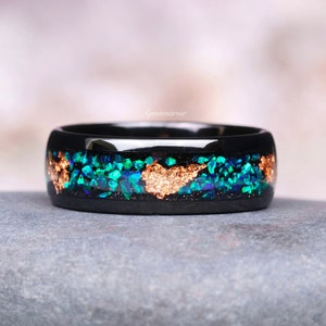 May include: A black ring with a band of crushed green and blue stones and gold leaf accents. The ring is set on a gray stone surface. The word "Aquamarise" is in the background.
