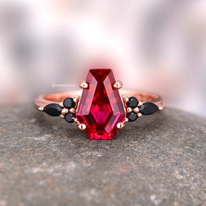 May include: A rose gold ring with a large, red, faceted gemstone in a coffin shape. The ring is accented with smaller black gemstones.