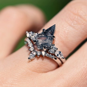 Pu&ograve; includere: Un set di anelli color argento con una grande pietra traslucida grigio scuro a forma di aquilone come elemento centrale, accentuato da pietre pi&ugrave; piccole, chiare, a taglio marquise e rotondo, e una fascia a forma di chevron con pietre chiare.